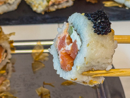 A person uses chopsticks to hold up a sushi roll filled with salmon, cream cheese, and topped with black caviar. The platter features several other sushi pieces, elegantly arranged.の写真素材