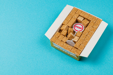 A yellow box filled with brown sugar cubes features a prominent stop sign. The bright blue background enhances the focus on the sugary contents, promoting mindful eating habits.の写真素材