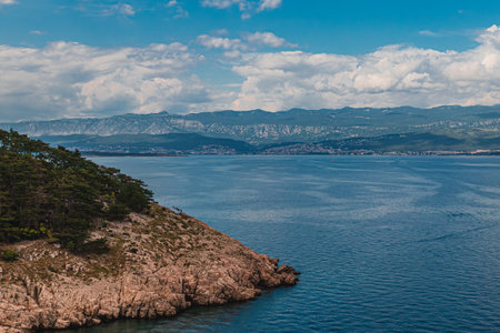 Serene waters gently lap against rugged cliffs, while lush greenery frames the coastline. Majestic mountains rise in the background under a vibrant sky, evoking tranquility.の写真素材