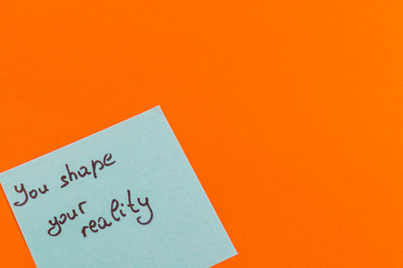 A handwritten note on a blue sticky pad encourages positivity with the phrase You shape your reality, placed against a bright orange background creating an inspiring atmosphere.の写真素材