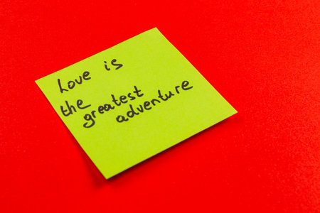 A bright green sticky note with a handwritten message stating love is the greatest adventure is placed on a vibrant red background. The note's colors create a striking visual contrast.の写真素材