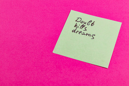 A green sticky note with the message Doubt kills dreams is placed on a bright pink background. This visual encourages self-confidence and positivity in daily life.の写真素材