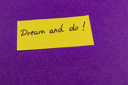 A bright yellow paper with the words Dream and do! handwritten on it is placed on a vibrant purple surface, conveying an inspiring motivational message.の写真素材