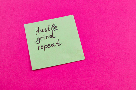 A green sticky note sits on a vivid pink surface, featuring the handwritten words hustle grind repeat. This simple reminder encourages persistence and hard work in daily life.の写真素材