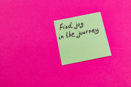 A green sticky note displays a motivational message encouraging individuals to find joy in their journey, set against a bright pink background that captures attention.の写真素材