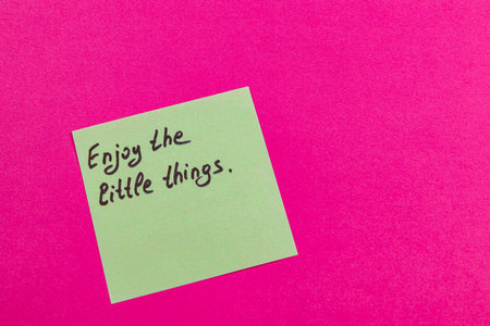 A green sticky note with a handwritten message promoting positivity and mindfulness is placed on a vibrant pink background. The note advises viewers to cherish small joys in life.の写真素材