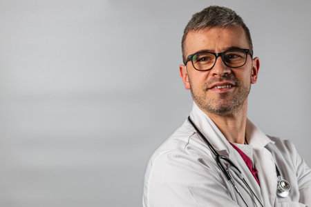 A healthcare professional wearing glasses and a white lab coat stands confidently, stethoscope draped around his neck, against a neutral gray backdrop, showcasing a caring demeanor.の写真素材