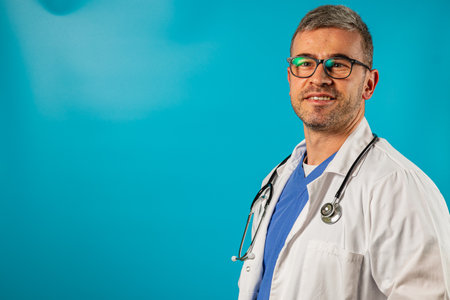 A healthcare professional stands confidently with a stethoscope around his neck. He wears a white coat over a blue scrub top and has a friendly smile. Bright blue backdrop enhances the atmosphere.の写真素材