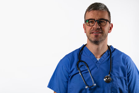 A male healthcare worker wearing blue scrubs and glasses stands confidently with a stethoscope around his neck. He smiles against a neutral background, ready for a professional setting.の写真素材