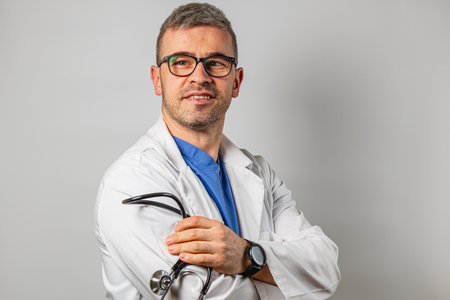 A male doctor smiles confidently while holding a stethoscope. Dressed in a white coat and blue scrubs, he conveys professionalism and approachability in a clinical setting.の写真素材