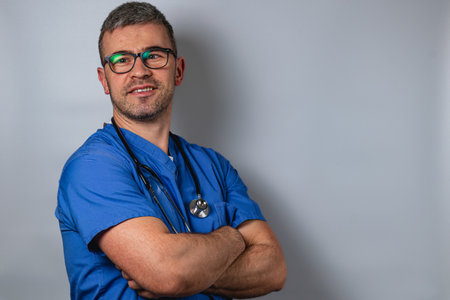 A healthcare professional wearing blue scrubs stands with arms crossed, showcasing a confident smile. The background is a solid gray, highlighting the individual's dedication to their work.の写真素材