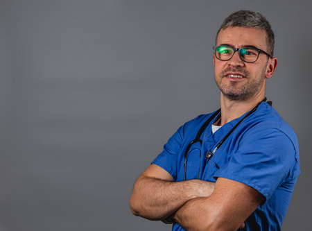 A healthcare worker stands with arms crossed, wearing blue scrubs and glasses. The setting shows a neutral background, emphasizing the dedication and professionalism of medical staff.の写真素材