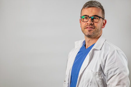 A male doctor wearing glasses and a white coat stands confidently with a slight smile, showcasing a blue scrub underneath, in a neutral setting.の写真素材