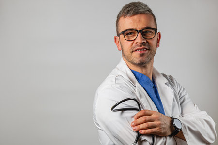A healthcare professional wearing a white coat and glasses stands with his arms crossed, holding a stethoscope. He looks confident and approachable, conveying professionalism in a clinical setting.の写真素材