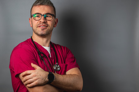 A male healthcare professional wearing scrubs and glasses stands with arms crossed against a neutral background, projecting confidence and professionalism in a medical setting.の写真素材