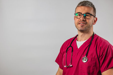 A male healthcare professional stands confidently, wearing red scrubs and a stethoscope around his neck. He has short grey hair and glasses, ready to assist patients in a medical setting.の写真素材