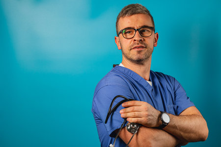 A male doctor wearing blue scrubs and glasses stands with his arms crossed, showcasing a stethoscope while looking thoughtfully in the distance against a vibrant blue background.の写真素材