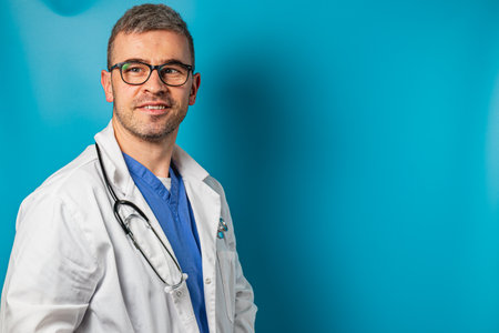 A healthcare professional stands with a slight smile, wearing a medical coat and scrubs. The bright blue background highlights his confident demeanor and professional appearance.の写真素材