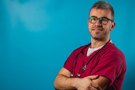 A healthcare professional wearing red scrubs and glasses stands confidently with arms crossed. The background is a vibrant blue, adding to the positive atmosphere of the moment.の写真素材