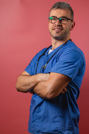 A healthcare professional stands with crossed arms, wearing blue scrubs and glasses. The vibrant backdrop enhances the confident demeanor, showcasing determination and competence in medicine.の写真素材