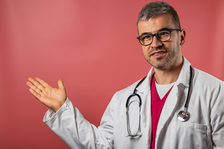 A doctor wearing a lab coat and glasses stands in a brightly lit room, smiling and gesturing with his right hand as if explaining something important to the viewer.の写真素材
