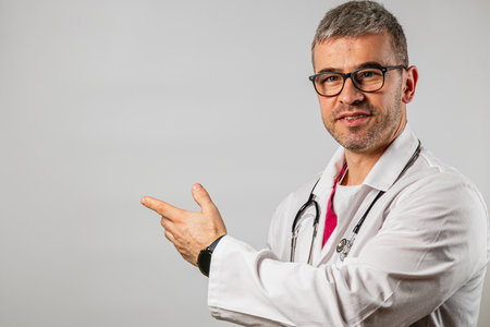 A doctor with glasses and a stethoscope gestures towards an empty space, indicating he is ready to provide information or guidance. The setting appears professional and clean.の写真素材