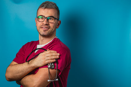 A male healthcare worker wearing pink scrubs and glasses stands confidently with his arms crossed. He smiles warmly while holding a stethoscope against a vibrant blue background.の写真素材