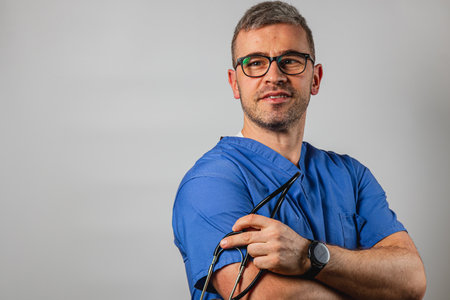A medical professional stands confidently wearing blue scrubs and holding glasses. The background is neutral, highlighting the subject's calm demeanor and readiness.の写真素材