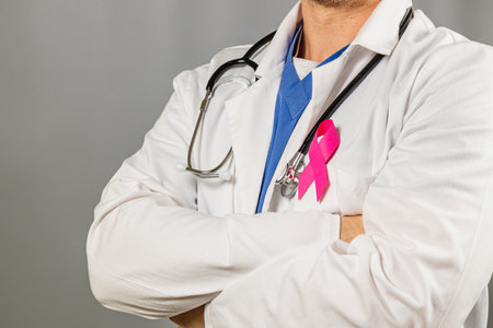 A healthcare professional stands with arms crossed, showcasing a pink ribbon on their lab coat, symbolizing support for breast cancer awareness. The setting conveys a message of solidarity.の写真素材
