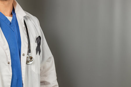 A healthcare professional stands wearing a white coat and stethoscope. The person has a blue scrub underneath and displays a black ribbon, symbolizing remembrance or mourning.の写真素材