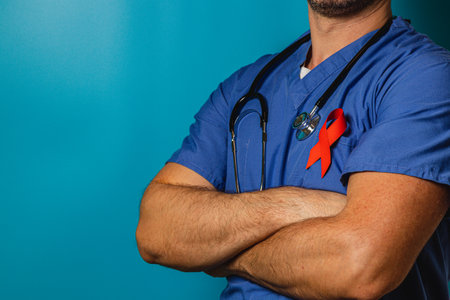 A healthcare worker stands confidently with arms crossed, wearing blue scrubs and a stethoscope, showcasing a red ribbon for HIV awareness against a solid blue background.の写真素材