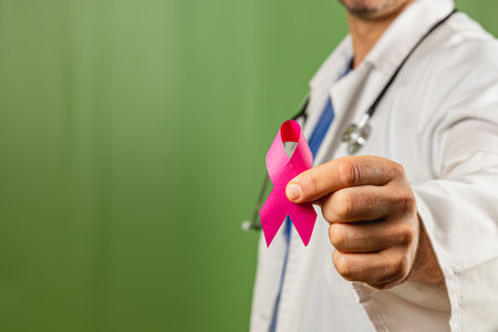 A doctor in a white coat holds a pink ribbon symbolizing breast cancer awareness. The backdrop is a green color, emphasizing the importance of early detection and support.の写真素材
