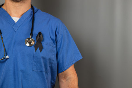 A healthcare professional in blue scrubs stands against a neutral backdrop. A black ribbon pinned on the chest symbolizes remembrance. The individual appears serious and reflective.の写真素材