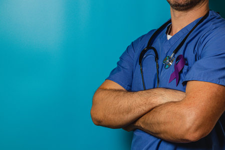 A healthcare professional stands confidently with arms crossed, wearing blue scrubs and a purple ribbon on his chest. The background is a solid teal color, creating a striking contrast.の写真素材