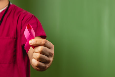 A healthcare worker in scrubs holds a pink ribbon to promote breast cancer awareness. The background features a simple green hue, emphasizing the importance of health education.の写真素材