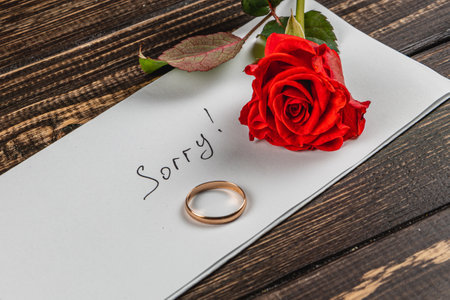 A heartfelt message written on a sheet of paper expresses sorrow for a broken engagement. A gold ring and a vibrant red rose accompany the note, resting on a wooden table.の写真素材