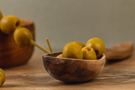 Green olives sit in a handcrafted wooden bowl, accompanied by toothpicks. The warm tones of the rustic table enhance the natural simplicity of the setting, inviting a taste experience.の写真素材