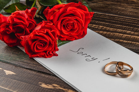 A note saying sorry is placed on a wooden surface alongside three red roses and two wedding rings, symbolizing sadness and remembrance of a love lost.の写真素材
