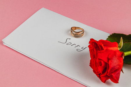 A gold engagement ring sits atop a white envelope with the word sorry handwritten on it. Beside the ring, a single red rose adds a touch of emotion, all against a soft pink backdrop.の写真素材