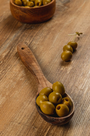 Green olives are arranged on a wooden table, with some in a serving spoon and others in wooden bowls. This rustic setting enhances their natural appeal.の写真素材