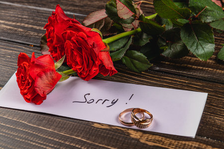 A note with the word Sorry lies beside two beautiful red roses and two wedding rings on a wooden table, expressing sadness over a broken engagement. The scene conveys deep emotional impact.の写真素材