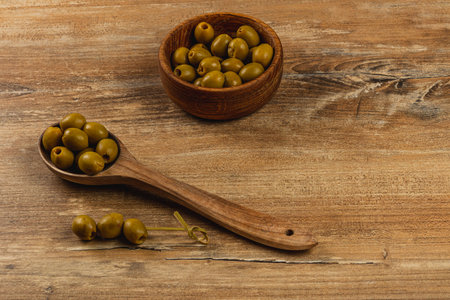 Green olives are arranged on a wooden surface along with a wooden spoon and a bowl. The rustic setting highlights the natural textures and colors of the ingredients.の写真素材