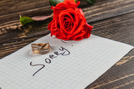 A note reading sorry is placed beside two wedding rings and a red rose on a rustic wooden surface, capturing the emotion of a broken engagement.の写真素材