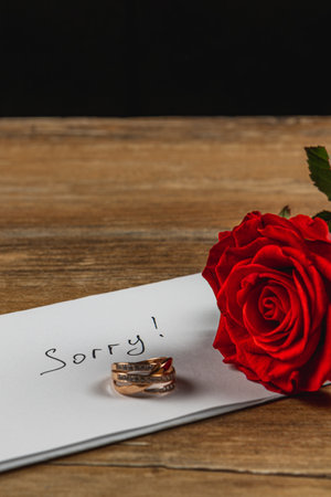 A note reading sorry lies on a wooden table next to two wedding rings and a red rose, capturing the emotions of a broken engagement in a simple yet poignant arrangement.の写真素材