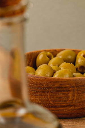 A rustic wooden bowl filled with fresh green olives sits on a table, while a glass bottle is softly blurred in the foreground, creating a warm ambiance in the setting.の写真素材