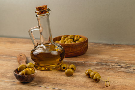 A glass bottle filled with olive oil sits alongside a wooden bowl of green olives. Some olives are scattered on the table, enhancing the rustic culinary theme.の写真素材
