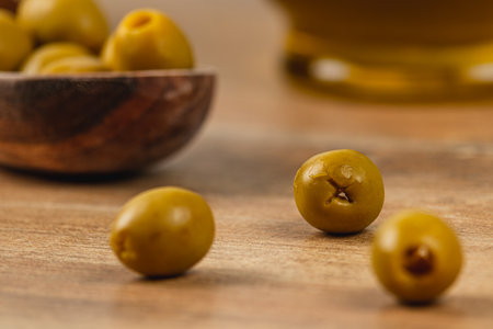 Fresh green olives are scattered on a wooden surface, accompanied by a rustic bowl and a glass of olive oil in the background, creating a warm and inviting atmosphere.の写真素材