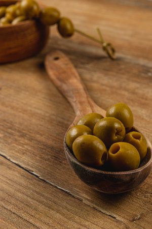 A wooden spoon holds several green olives on a rustic wooden table. Nearby, a bowl with more olives adds to the natural setting, highlighting fresh ingredients and culinary focus.の写真素材