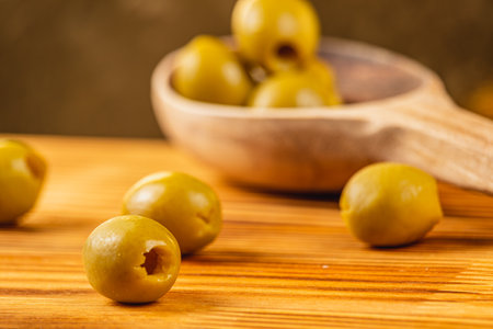 Juicy green olives are scattered across a wooden table, with a wooden spoon in the background holding more olives. The warm tones create a rustic and inviting atmosphere.の写真素材