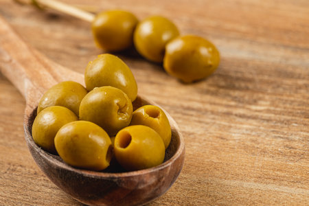 Several green olives are carefully placed on a wooden spoon. The spoon rests on a rustic wooden table, creating a warm atmosphere.の写真素材